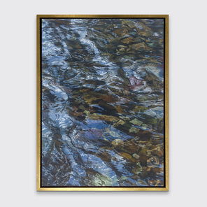 A brown, dark green, white and light blue abstract river rocks in water print in a gold floater frame hangs on a white wall.