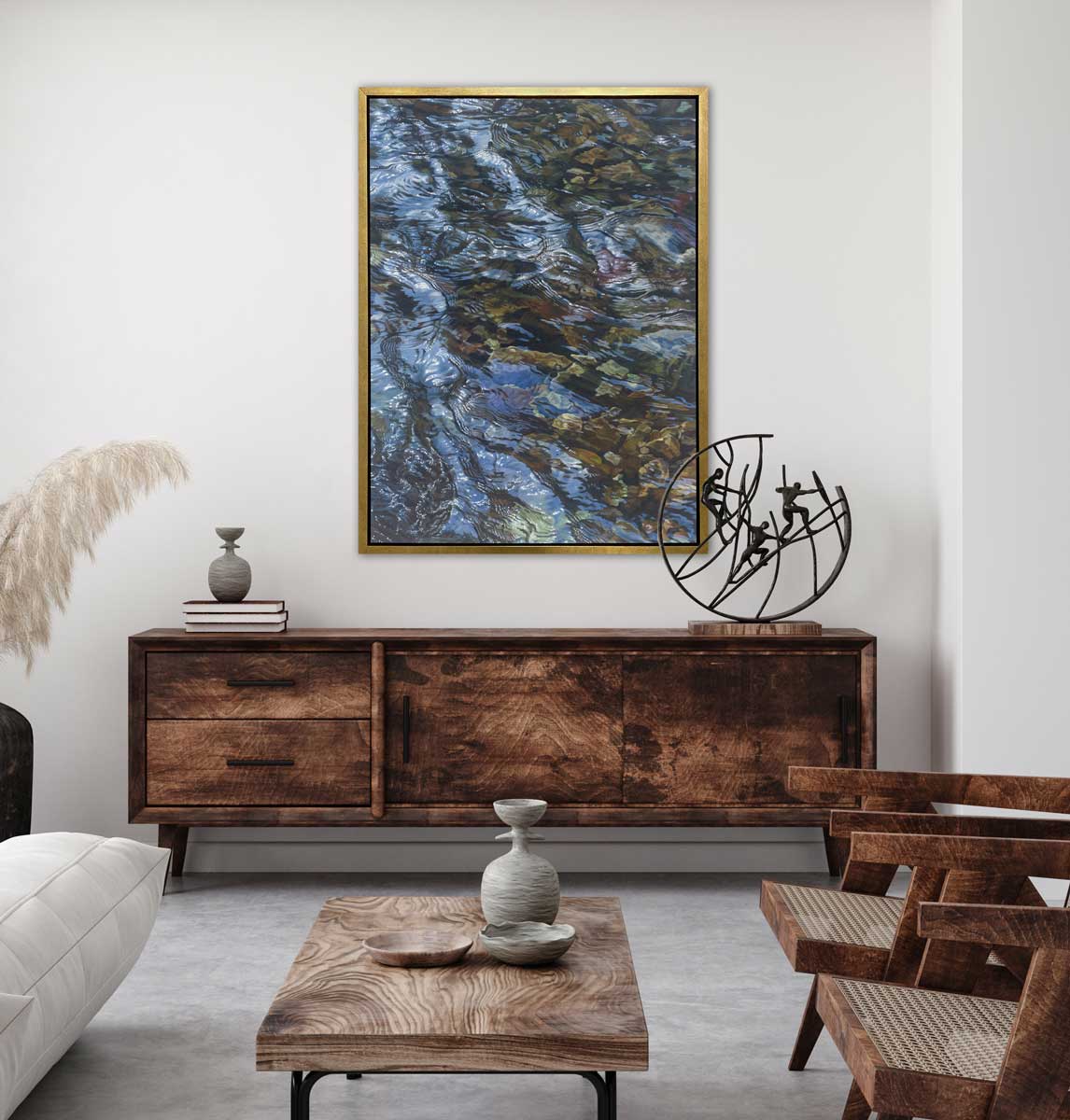 A brown, dark green, white and light blue abstract river rocks in water print in a gold floater frame hangs on a white wall above a dark wood stained console cabinet with accessories, two wood and rattan chairs and a rectangular wood coffee table.