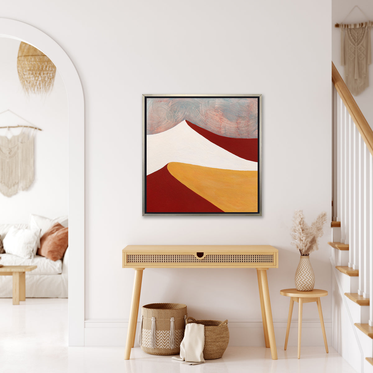 A dark red, white, dark yellow and teal abstract landscape print in a silver floater frame hangs on a white wall above a natural wood console desk with a drawer, two woven baskets and a small side table with a wheat filled vase.