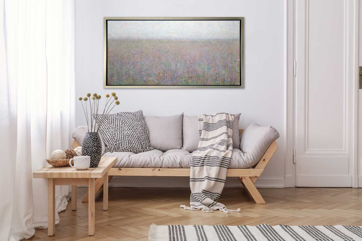 A green and purple abstract landscape print in a silver floater frame hangs on a white wall above a wooden sofa with grey cushions, a black and white printed pillow and a black and white striped throw blanket and a wooden side table with accessories.