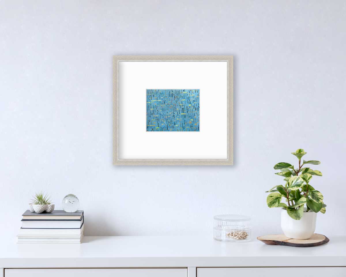 A blue, yellow and orange abstract print in a silver frame with a mat hangs on a white wall above a white surface with a stack of books, a round clear container with gold accessories and a potted plant.