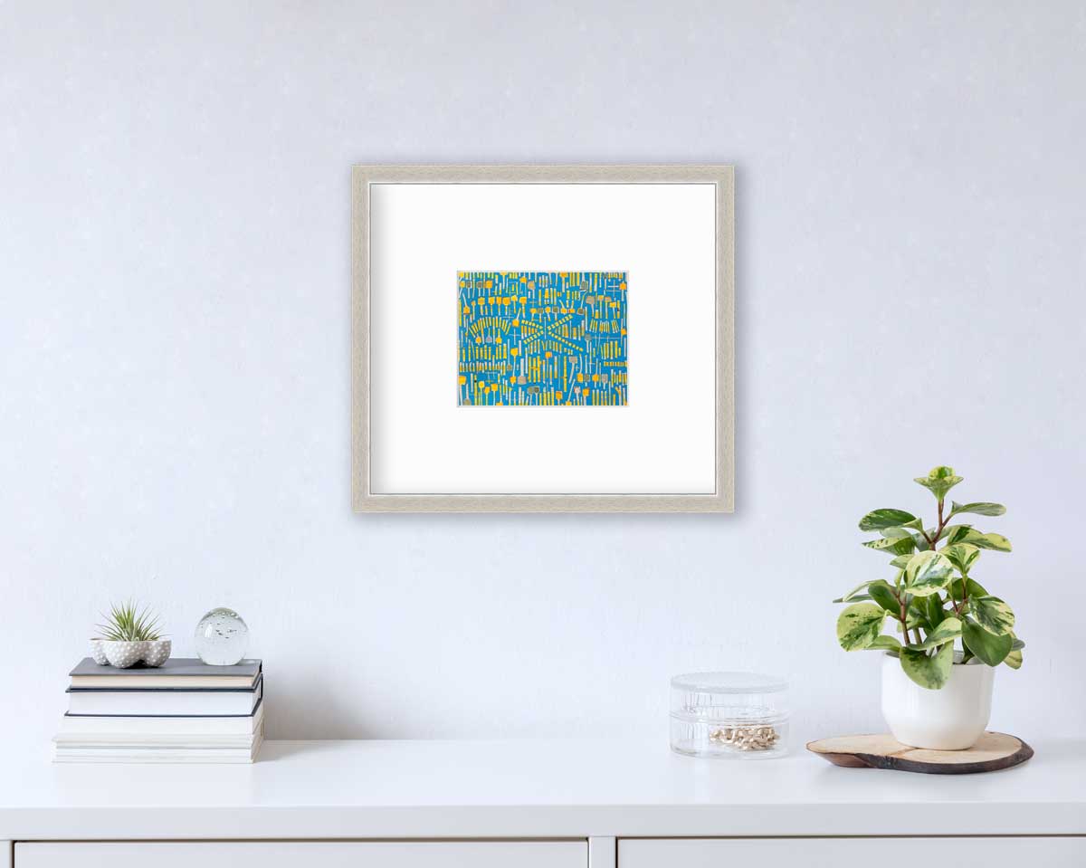 A blue and yellow abstract print in a silver frame with a mat hangs on a white wall above a white surface with a stack of books, a round clear container with gold accessories and a potted plant.