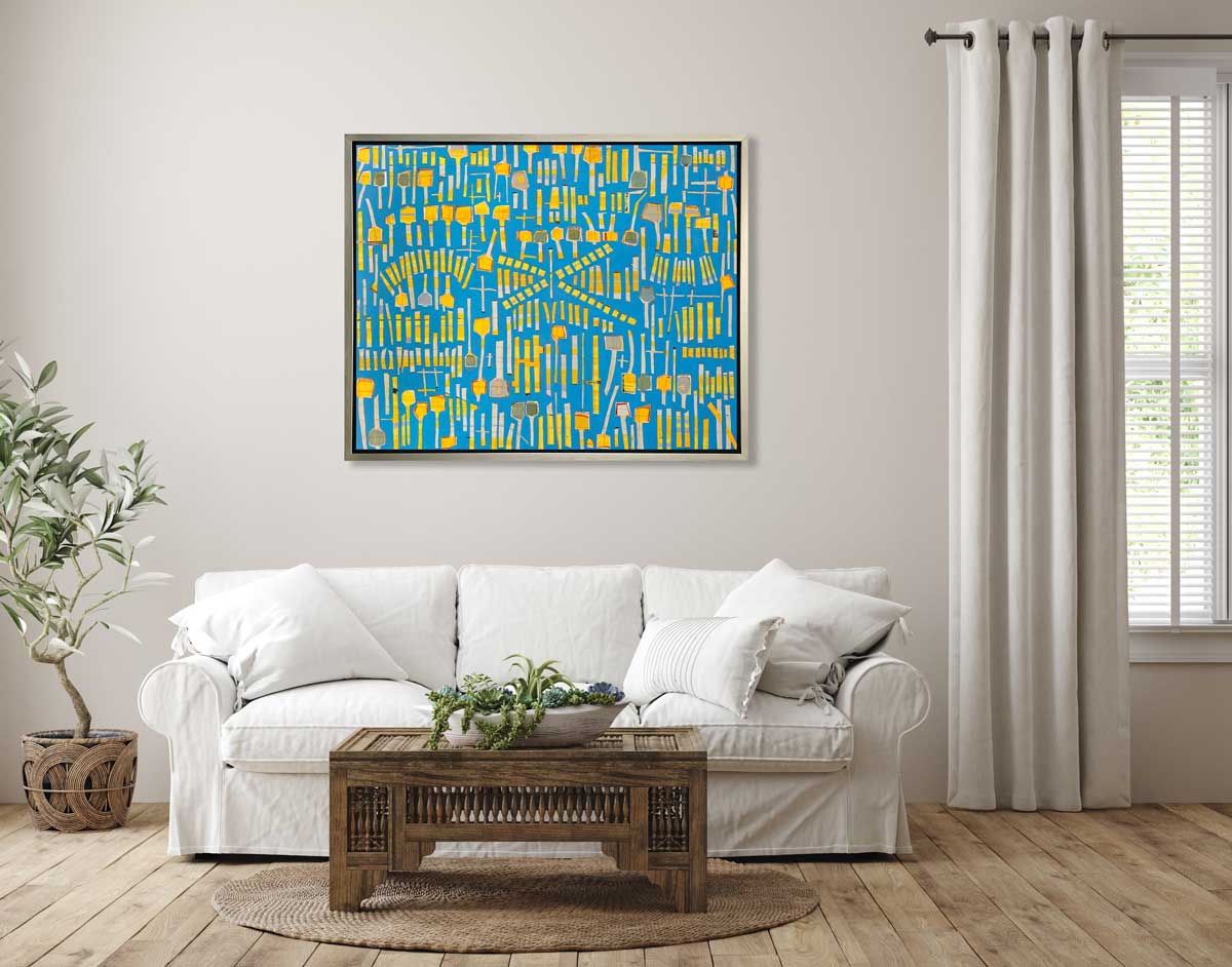 A blue and yellow abstract print in a silver floater frame hangs on a white wall above a white sofa with pillows, an ornate wood coffee table with a succulent arrangement and a potted tree.