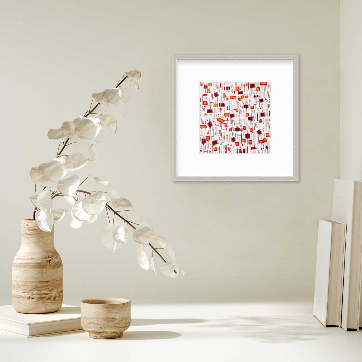A red, white and orange abstract print in a silver frame with a mat hangs on a white wall above a stack of books and a wooden vase with white leaf stems.