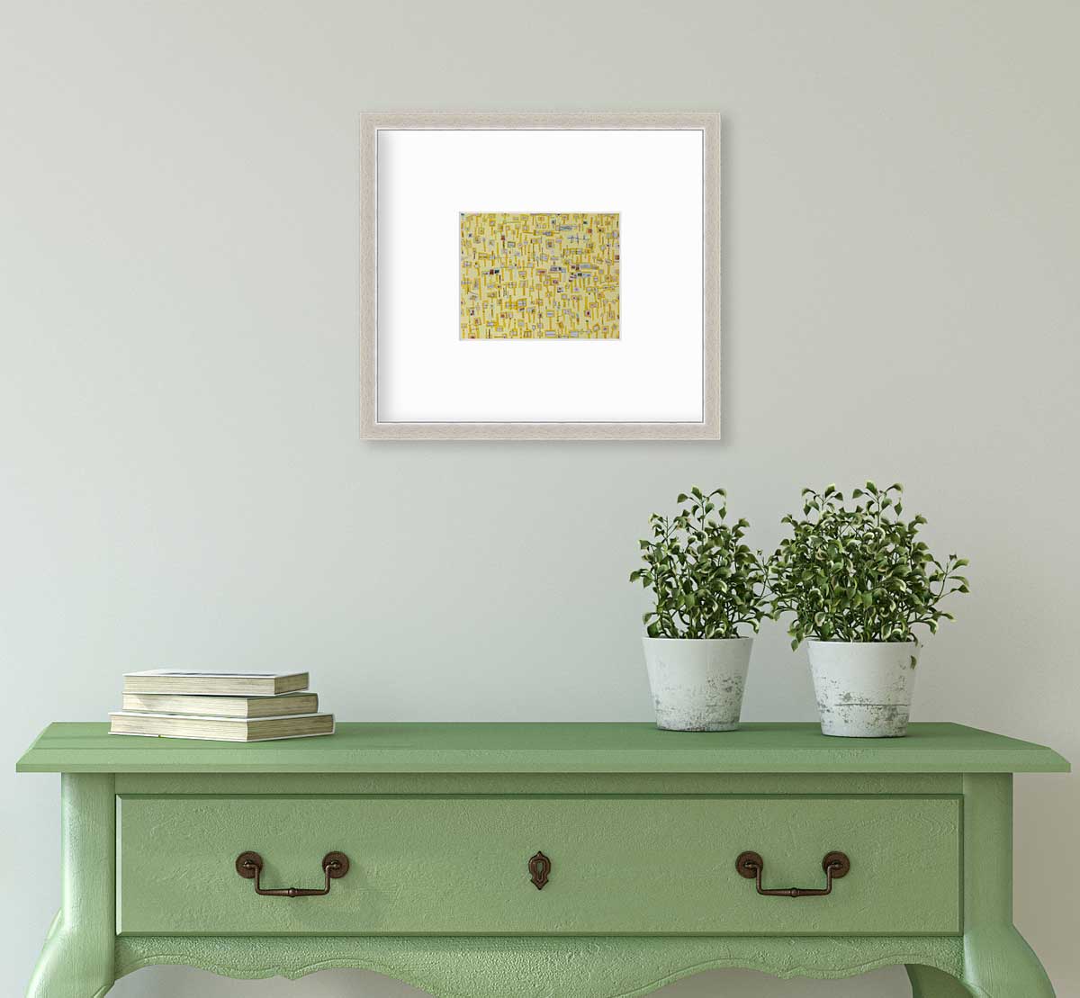 A yellow and grey abstract print in a silver frame with a mat hangs on a white wall above a green console table with a stack of books and two potted plants.