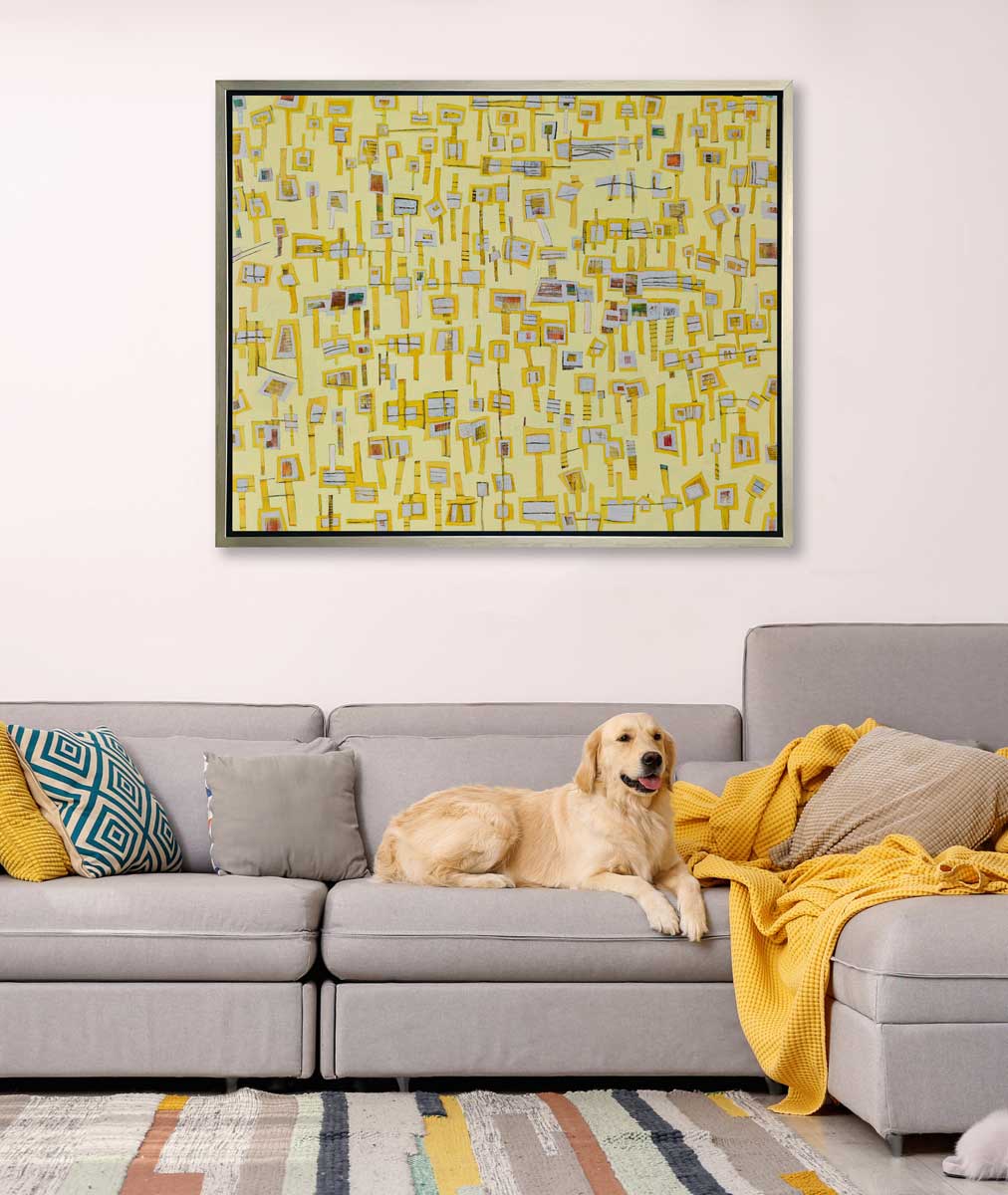 A yellow and grey abstract print in a silver floater frame hangs on a white wall above a grey sofa with grey pillows, a yellow throw blanket and a yellow Labrador Retriever laying down.