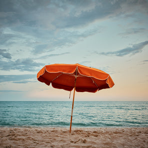 A photograph of an orange umbrella on a sandy beach at sunset. 