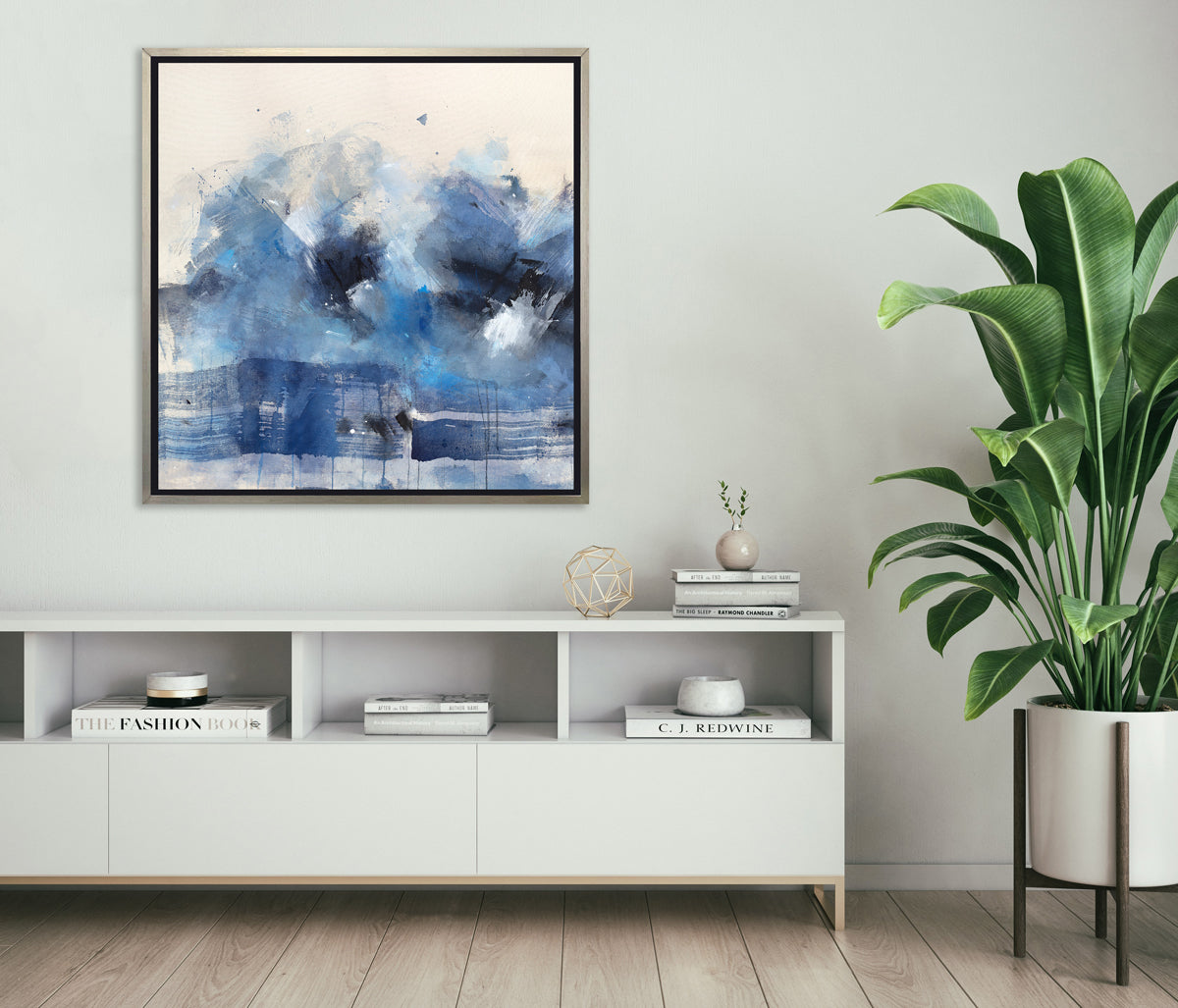 A blue, beige, white and black abstract print in a silver floater frame hangs on a white wall above a white storage cabinet with stacks of books, a gold wire sphere and a large potted floor plant to the right.