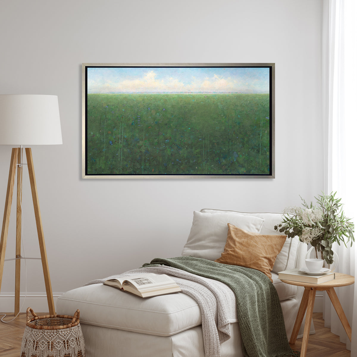 A green, light blue and white abstract landscape print in a silver floater frame hangs on a white wall above a white lounger chair with pillows and throw blankets, a small round natural wood side table and a tall tripod floor lamp.