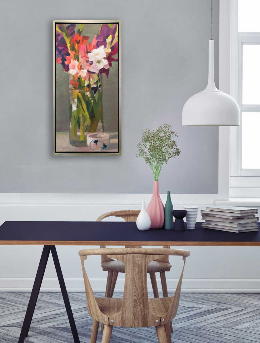 A purple, pink and green abstract floral print in a silver floater frame hangs on a grey wall above a blue table with a stack of books, several vases in different colors and two wood chairs.