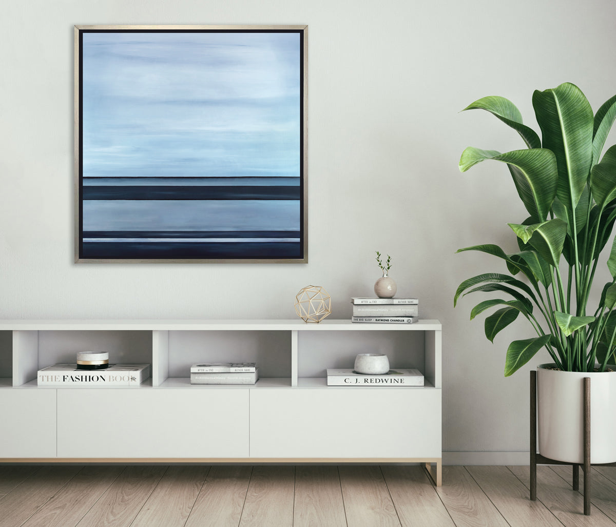 A tonal blue linear abstract print in a silver floater frame hangs on a white wall above a white credenza with stacks of books, a gold geometric wire sphere and a tall potted floor plant.