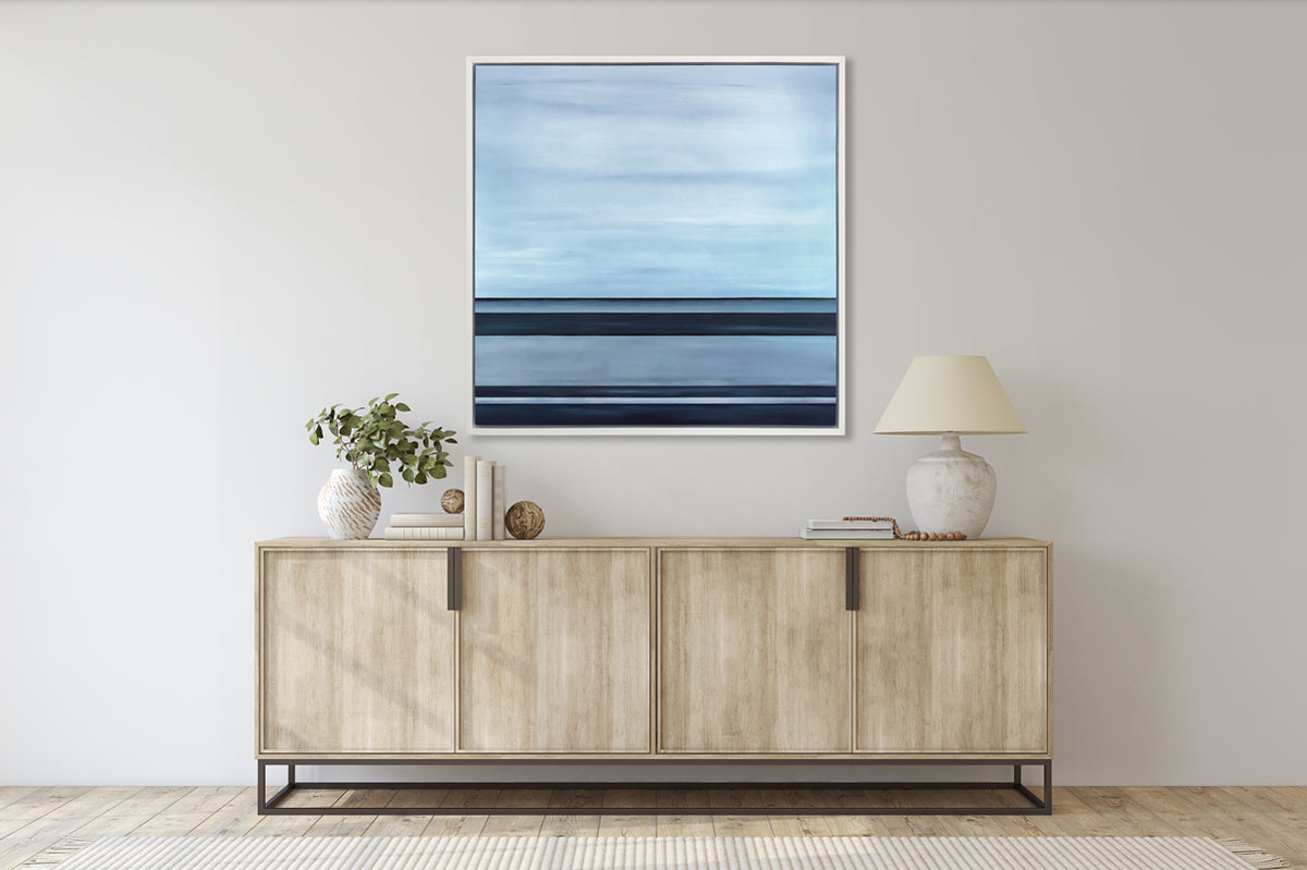 A tonal blue linear abstract print in a white floater frame hangs on a white wall above a natural wood credenza with a white table lamp, stacks of books, two decorative spheres and a eucalyptus filled white and terracotta vase.
