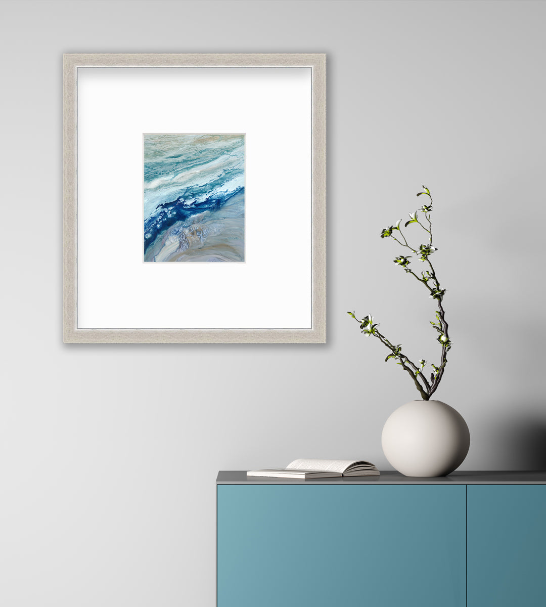 A blue, teal, gold and white abstract print in a silver frame with a mat hangs on a white wall above a teal cabinet with an open book and a round white vase with greenery stems.