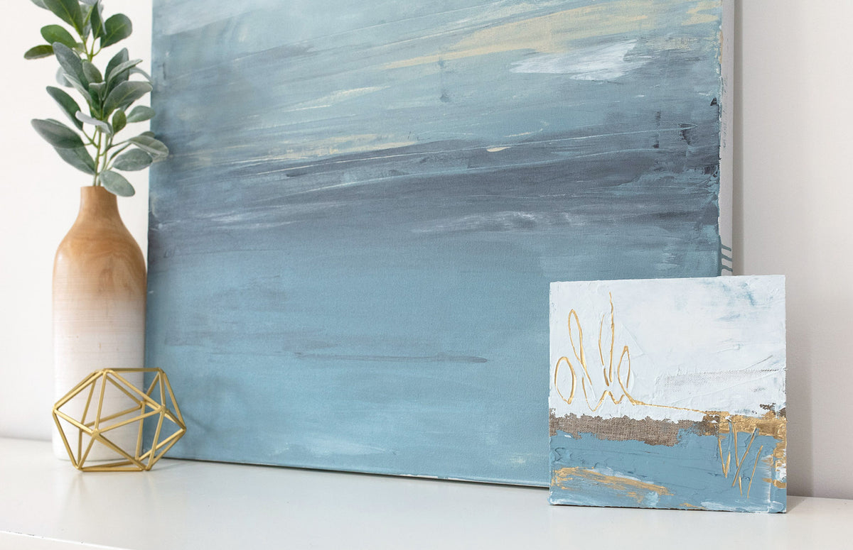 Two abstract paintings by Julia Contacessi resting on a white dresser next to modern table decor. 