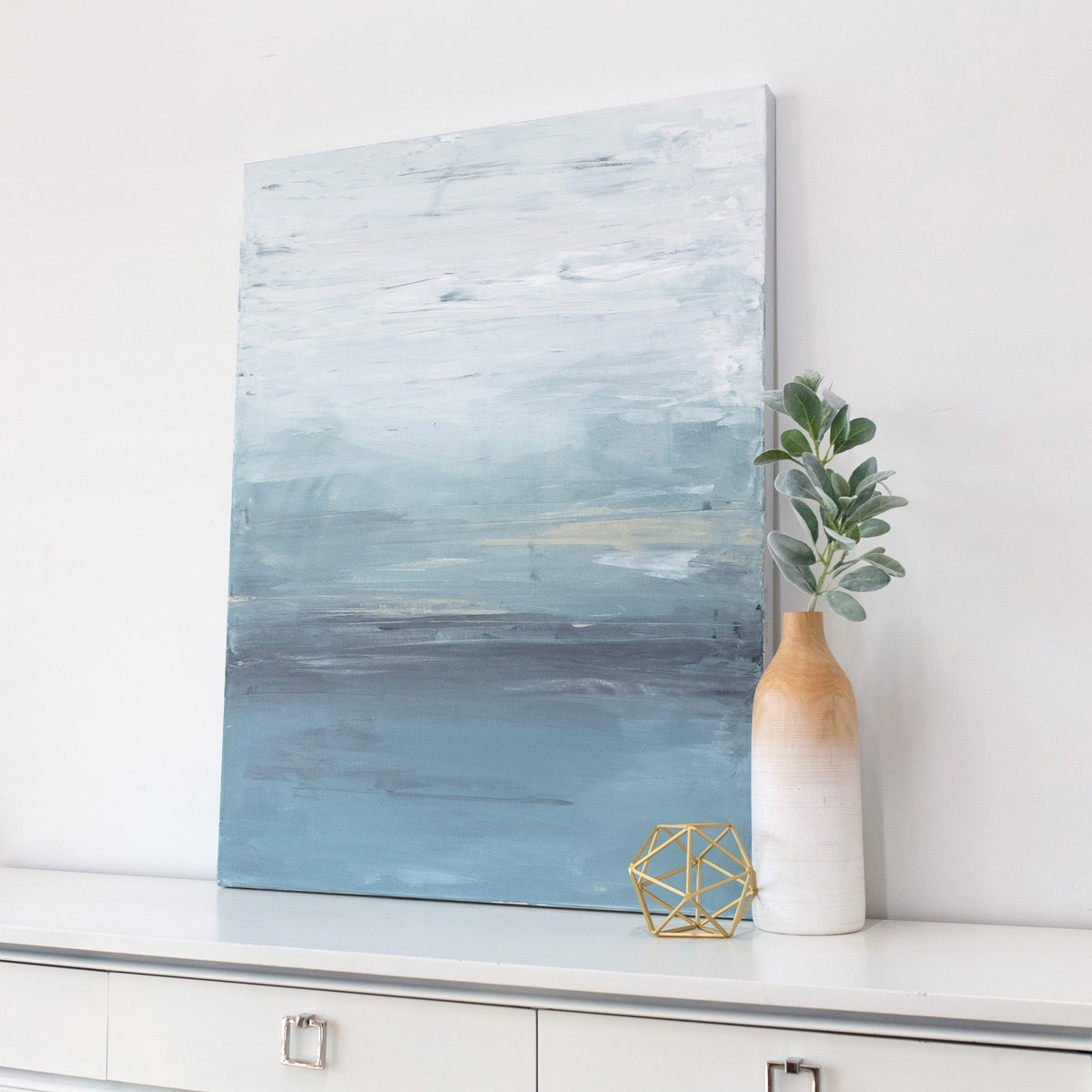 A side-angle view of a blue abstract painting resting on a modern white dresser next to modern table decor. 