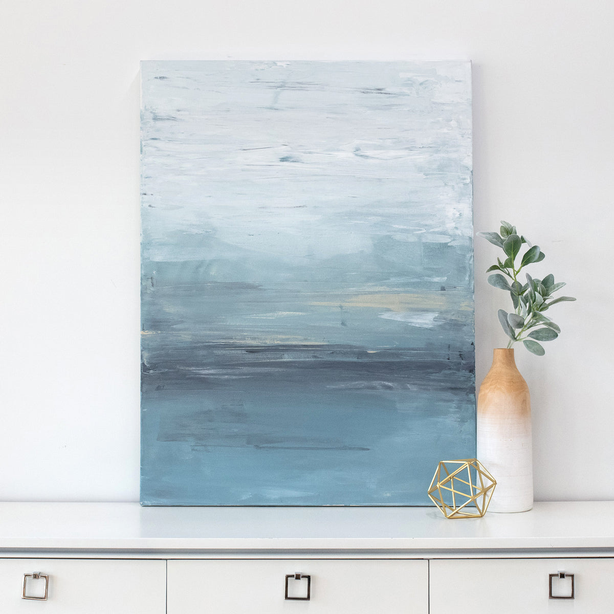 A blue abstract painting rests on a white dresser next to modern table decor. 