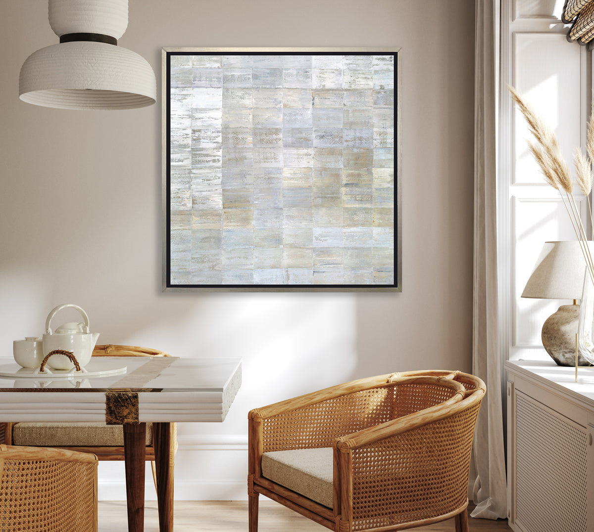 A grey, silver and light gold abstract geometric print in a silver floater frame hangs on a white wall above a glass top and wood table, three rattan chairs with cushions and a white window shelf with a white rock table lamp.