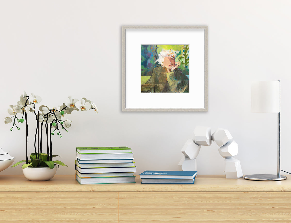 A green, beige and white abstract figural print in a silver frame with a mat hangs on a white wall above a natural wood shelf with stacks of books, a potted white orchid, a white blocked arch decoration and a small modern lamp.