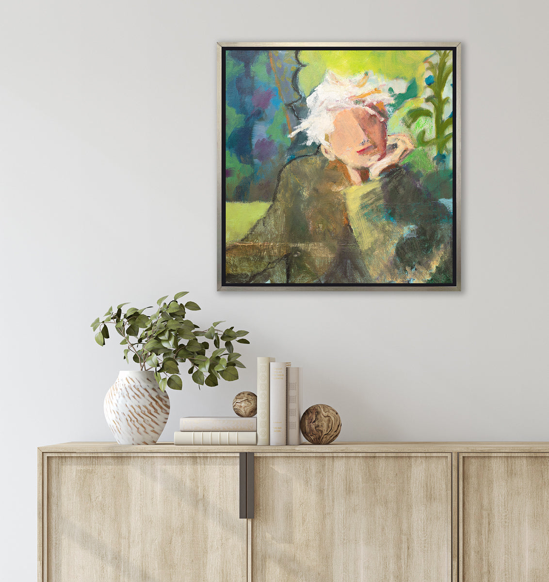 A green, beige and white abstract figural print in a silver floater frame hangs on a white wall above a natural wood credenza with a stack of books, two wood sphere decorations and a white vase with eucalyptus leaves.