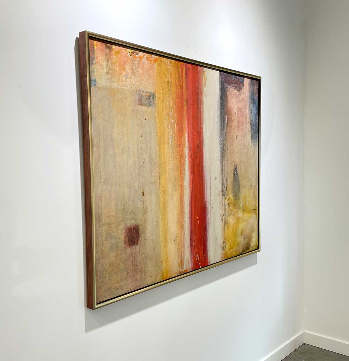 A left side-angle view of a multicolored abstract painting in a warm gold frame hangs on a white wall.