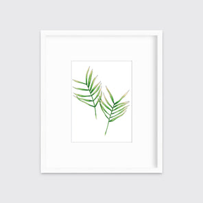 A print of a two green leaves with stems with hints of yellow in a white frame with a mat hangs on a white wall. 