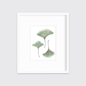 A print of three green leaves from a maidenhair fern with hints of yellow and brown in a white frame with a mat hangs on a white wall. 