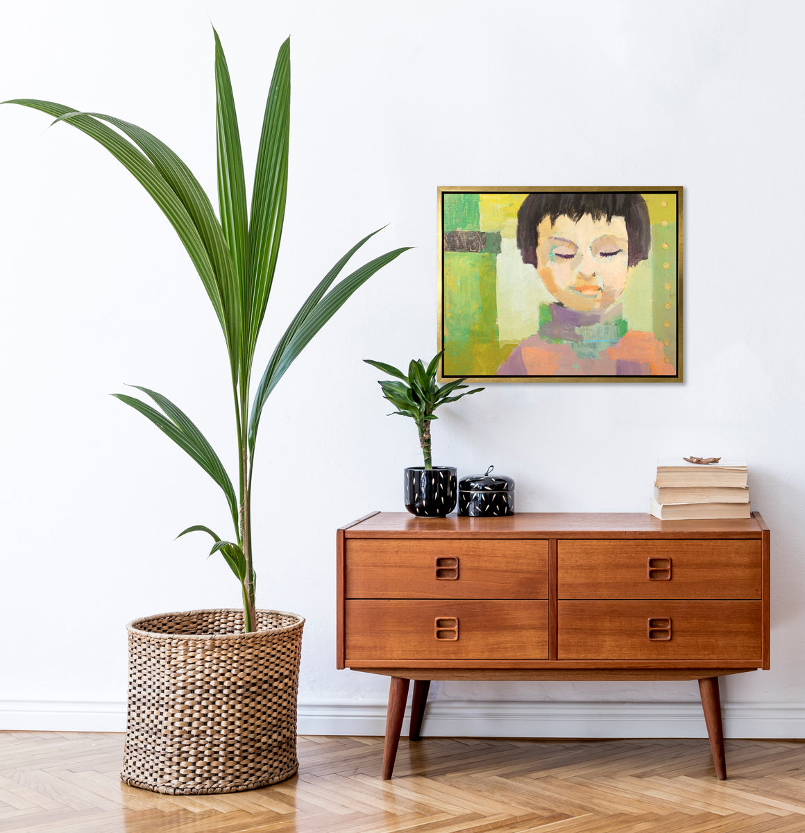 A green, coral, beige and brown abstract figural print in a gold floater frame hangs on a white wall above a stained wood cabinet with a stack of books, a small potted plant, a black container with a lid and a large potted floor plant.