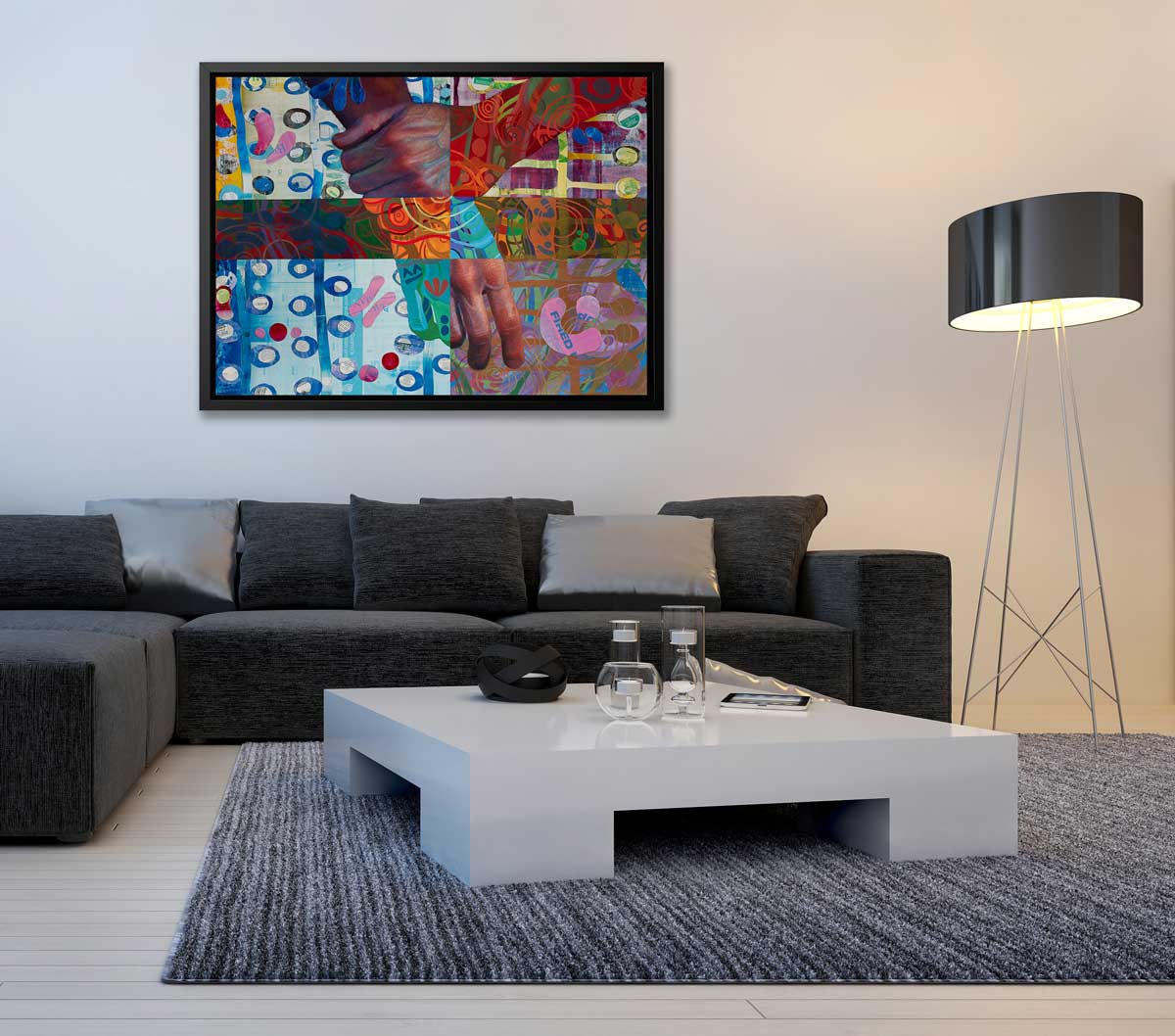 A multicolored abstract print of two hands in a black floater frame hangs on a white wall above a black sofa with black and silver pillows, a light grey square coffee table and a tall floor lamp with a black shade.