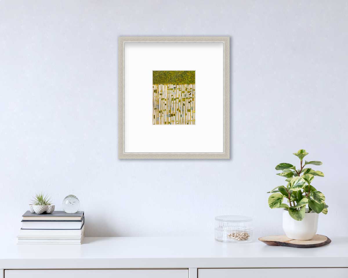 A green, yellow and white abstract print in a silver frame with a mat hangs on a white wall above a white surface with a stack of books, a round clear container with gold accessories and a potted plant.