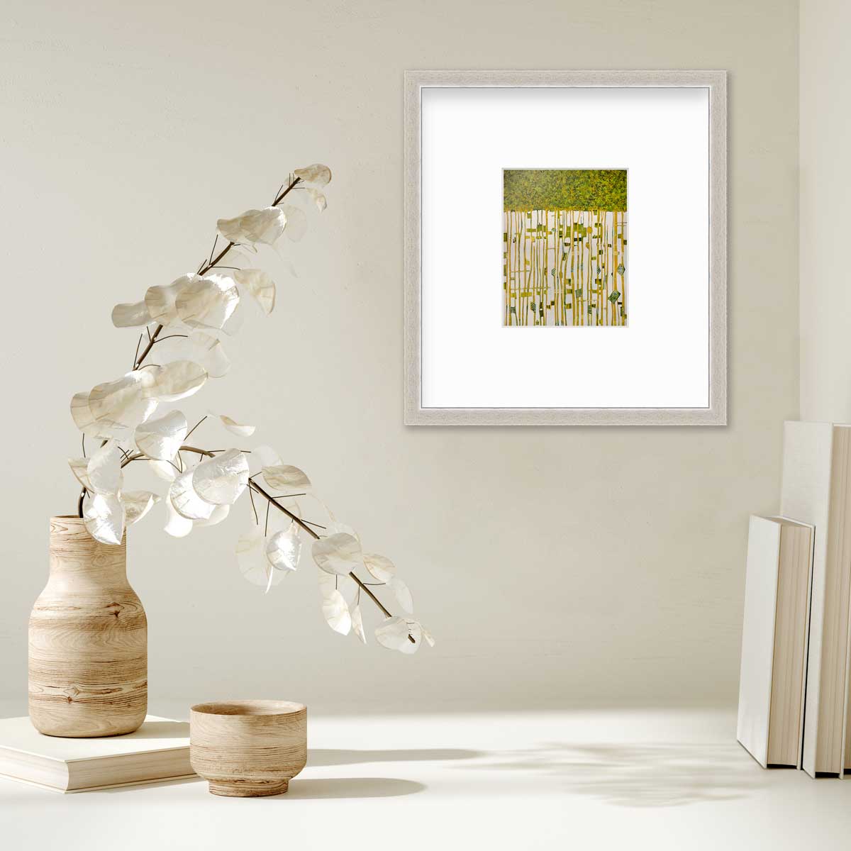 A green, yellow and white abstract print in a silver frame with a mat hangs on a white wall above a stack of books and a wooden vase with white leaf stems.