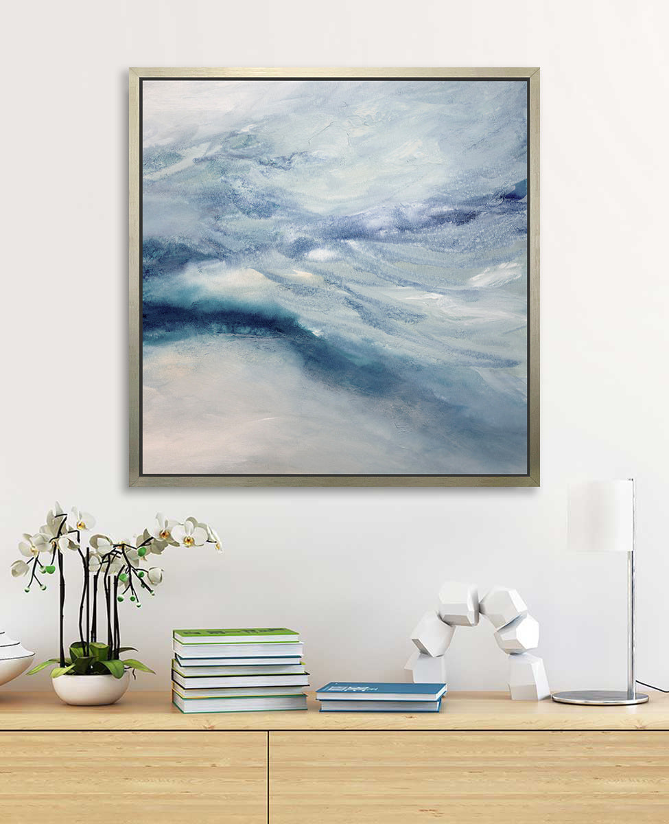 A blue, white, teal and light purple abstract print in a silver floater frame hangs on a white wall above a wood shelf with stacks of books, a white block decoration, a white and chrome desk lamp and a potted white orchid.