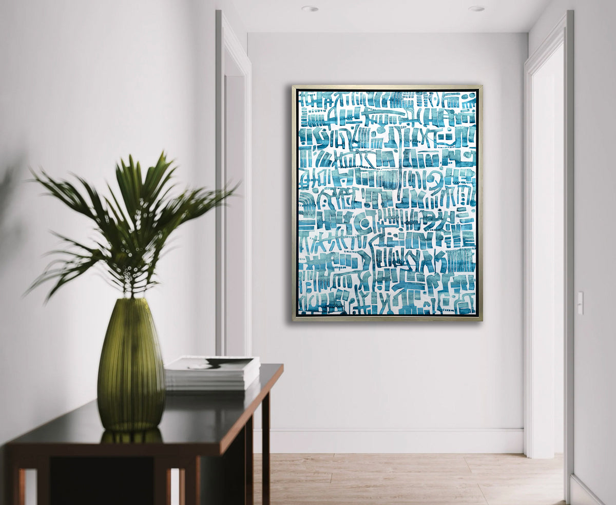 A teal and white abstract shapes print in a silver floater frame hangs on a white wall at the end of a hallway across from a black console table with a stack of books and a green glass vase with palm leaves.