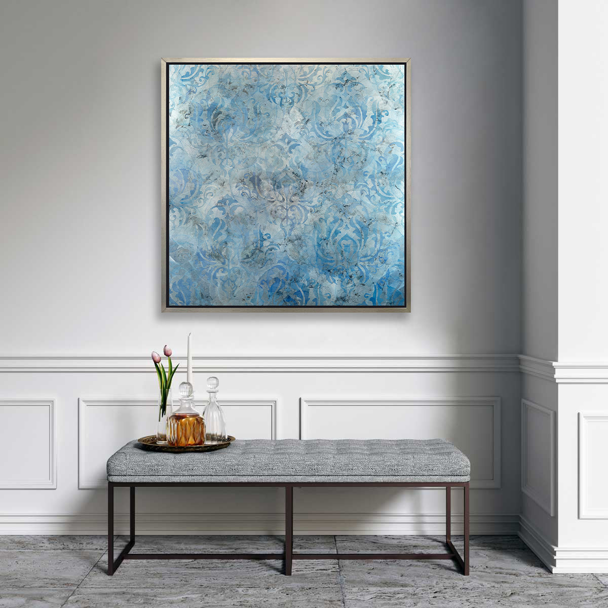 A dark blue, light blue and silver abstract print in a silver floater frame hangs on a white wall above a grey tufted bench and a tray with two glass decanters, a white tapered candlestick and a clear vase with two pink tulips.