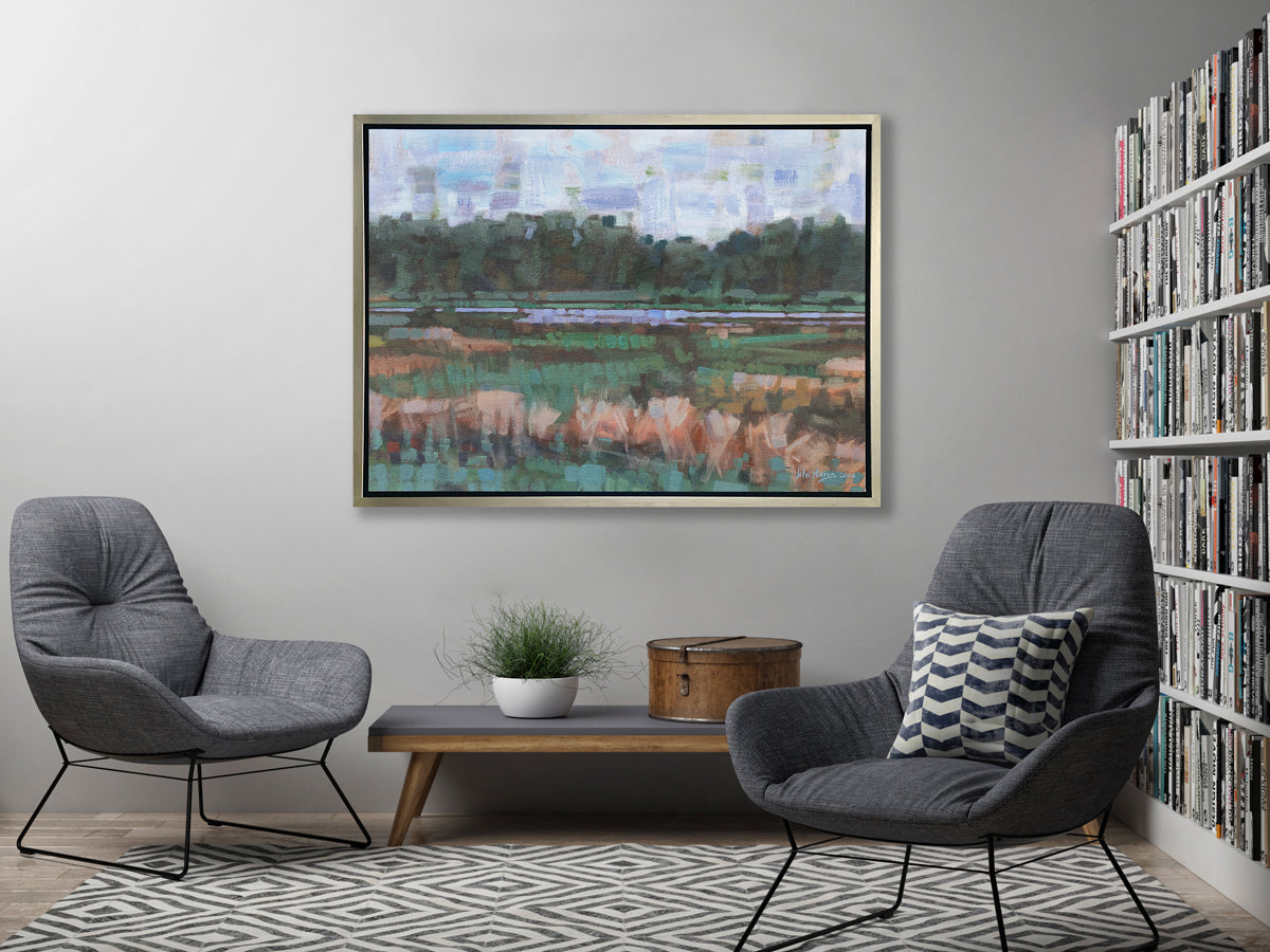 A green, orange and blue abstract landscape print in a silver floater frame hangs on a grey wall above a grey bench with a potted plant and a round hat box, two matching grey chairs and next to a large bookshelf.