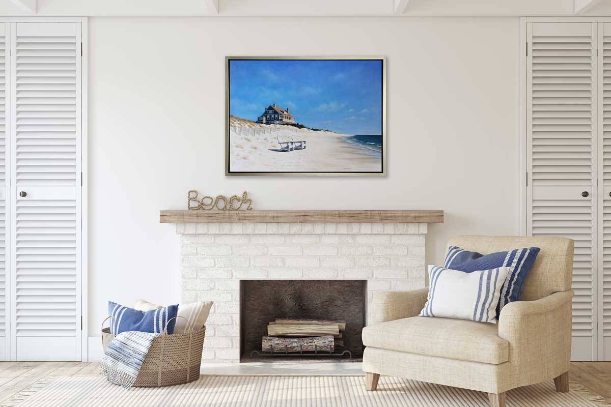 A contemporary realist seascape print in a silver floater frame hangs on a white wall above a white brick fireplace, a linen chair with blue and white striped pillows and a woven basket with more pillows.