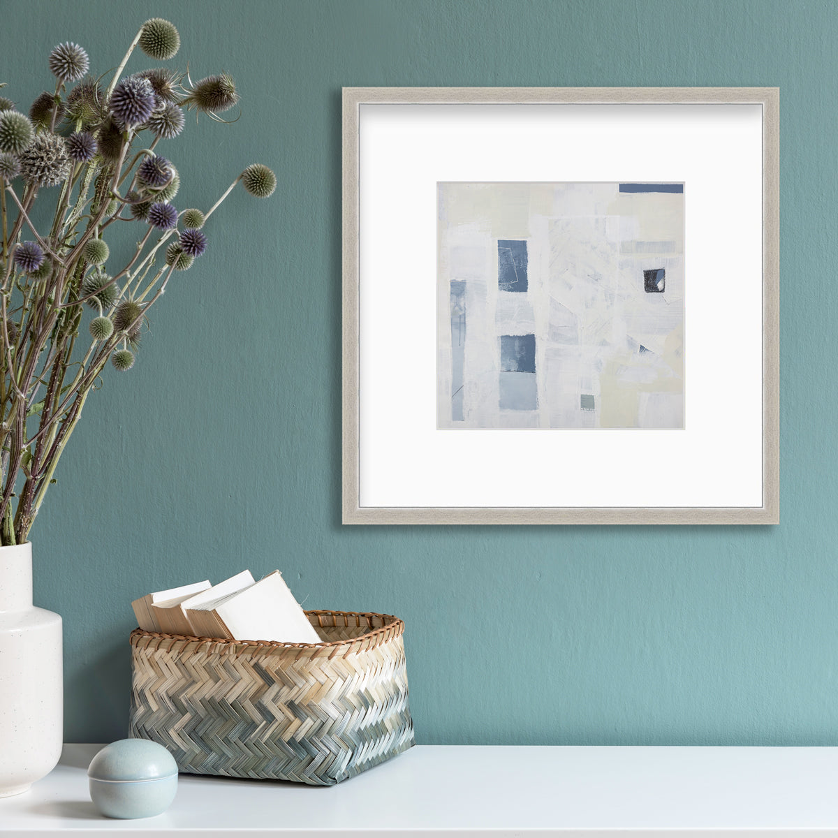 A white, pale yellow and slate blue abstract print in a silver frame with a mat hangs on a teal wall above a white surface with a small teal decoration, a teal and natural woven basket with card stock and a white vase with purple and green pom pom floral stems.