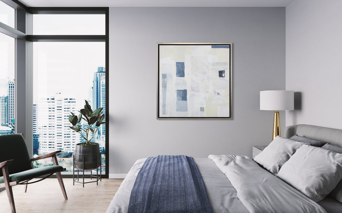 A white, pale yellow and slate blue abstract print in a silver floater frame hangs on a light grey wall near a large bed with grey bedding and a slate blue throw blanket, a modern chair and potted plant sit in the corner full of large floor to ceiling windows.