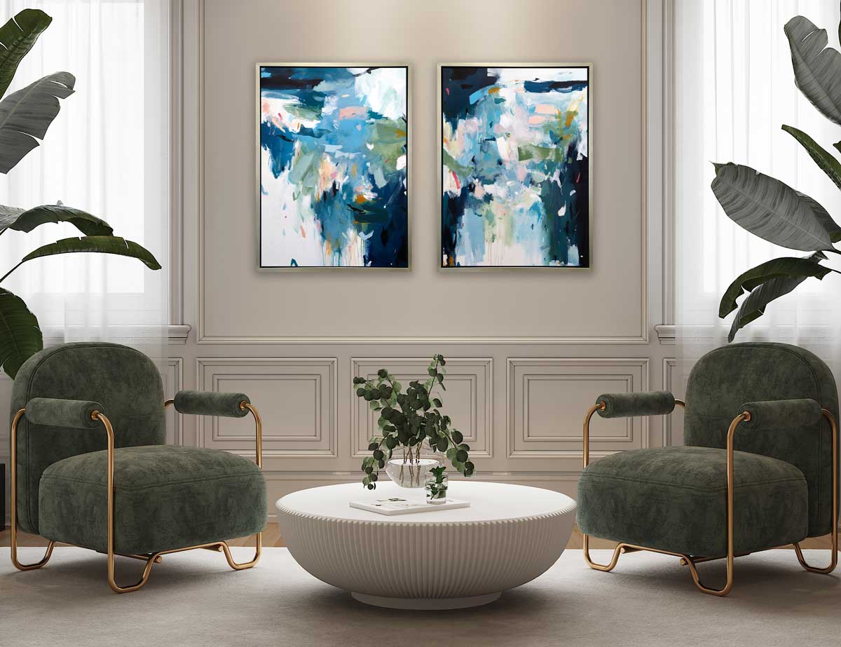 Two blue abstract prints in silver floater frames hang on a cream wall above two dark sage contemporary chairs with gold accents and a white coffee table.