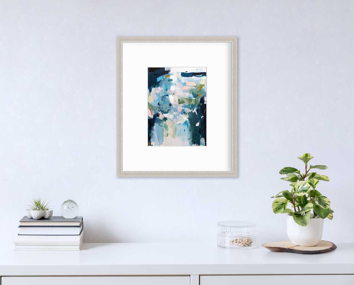 A blue abstract print by Kelly Rossetti in a silver frame with a mat hangs on a white wall above a white surface with a stack of books, a round clear container with gold accessories and a potted plant.