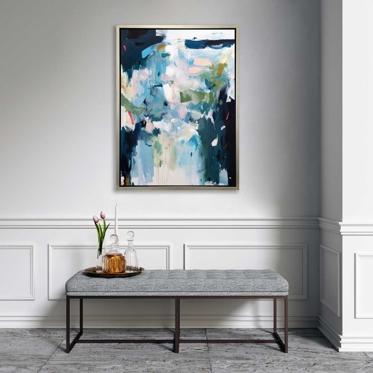 A blue, white and green abstract print in a silver floater frame hangs on a white wall above a grey tufted bench with a tray of two glass decanters, a tapered white candle and a glass vase with pink tulips.