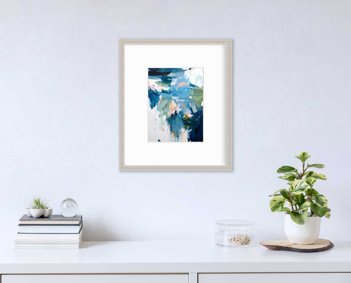 A blue abstract print by Kelly Rossetti in a silver frame with a mat hangs on a white wall above a white surface with a stack of books, a round clear container with gold accessories and a potted plant.