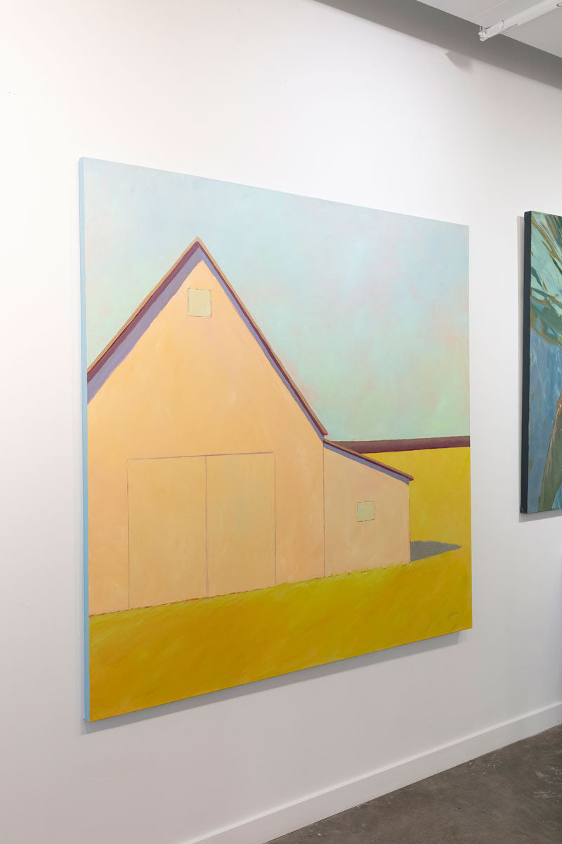 A painting on a gallery wall is seen at an angle. In the painting a large, cream-colored barn sits in a yellow field. Wired and ready to hang.