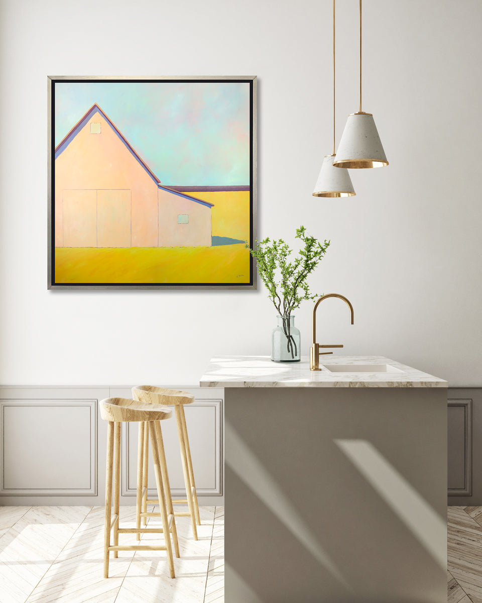A light blue, coral and deep yellow landscape print of a barn in a silver floater frame hangs on a white wall above a grey and white marble kitchen island and two natural wood stools.