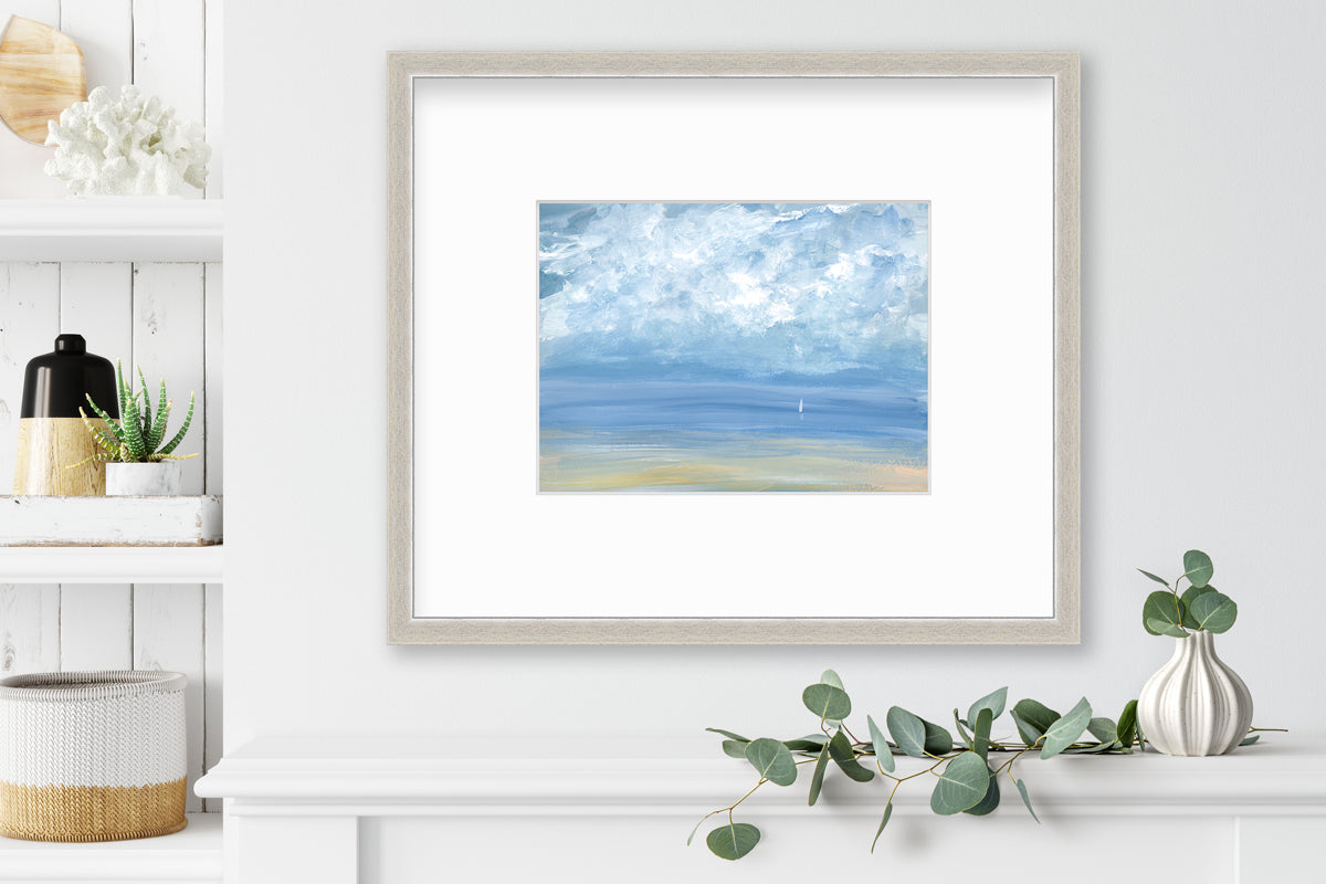 A white, blue and beige abstract seascape print in a silver frame with a mat hangs on a white wall above a white shelf with a eucalyptus filled white vase and next to a built in bookshelf with decorative accessories.