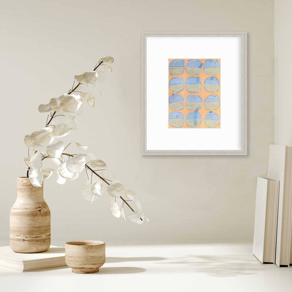 A orange, blue, green and white abstract print in a silver frame with a mat hangs on a white wall above a stack of books and a wooden vase with white leaf stems.