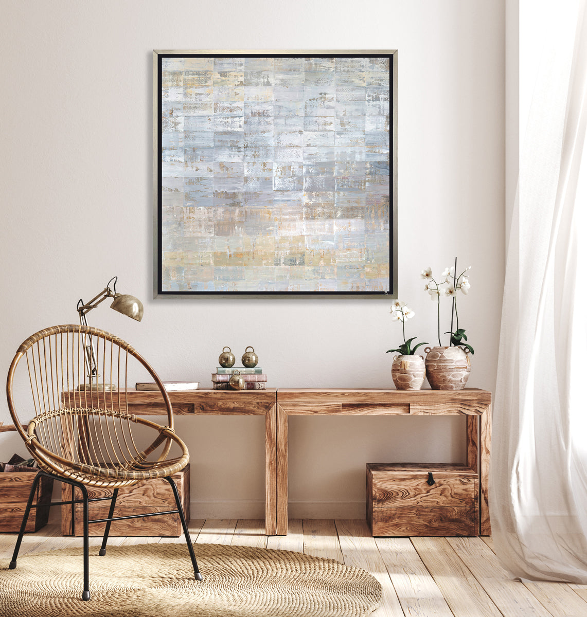 A grey, silver and light gold abstract geometric print in a silver floater frame hangs on a white wall above a long wood desk with two potted white orchids, a stack of books, a chrome adjustable desk lamp and a rounded rope chair.