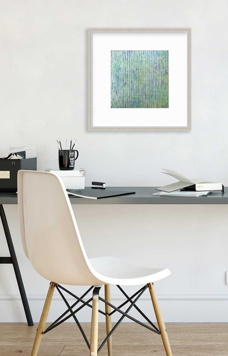 A green, blue and yellow abstract print in a silver frame with a mat hangs on a white wall above a grey desk with a black mug with pencils, open books and notebooks and a white chair.