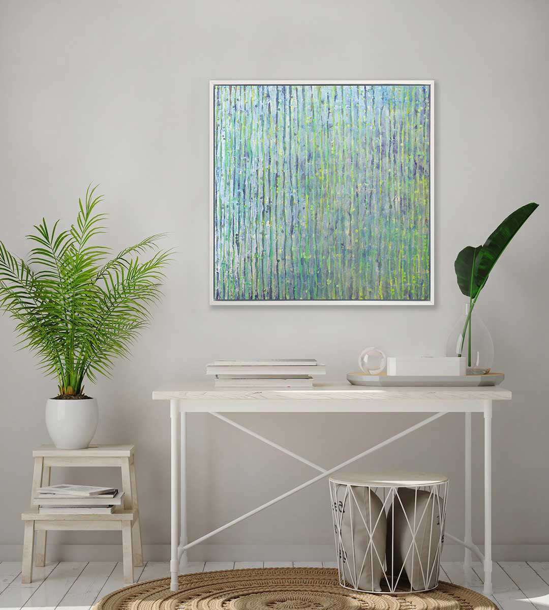A green, blue and yellow abstract print in a white floater frame hangs on a white wall above a white desk with stacked books and a tray, a small step stool on the floor with a potted plant and a small basket with pillows inside.