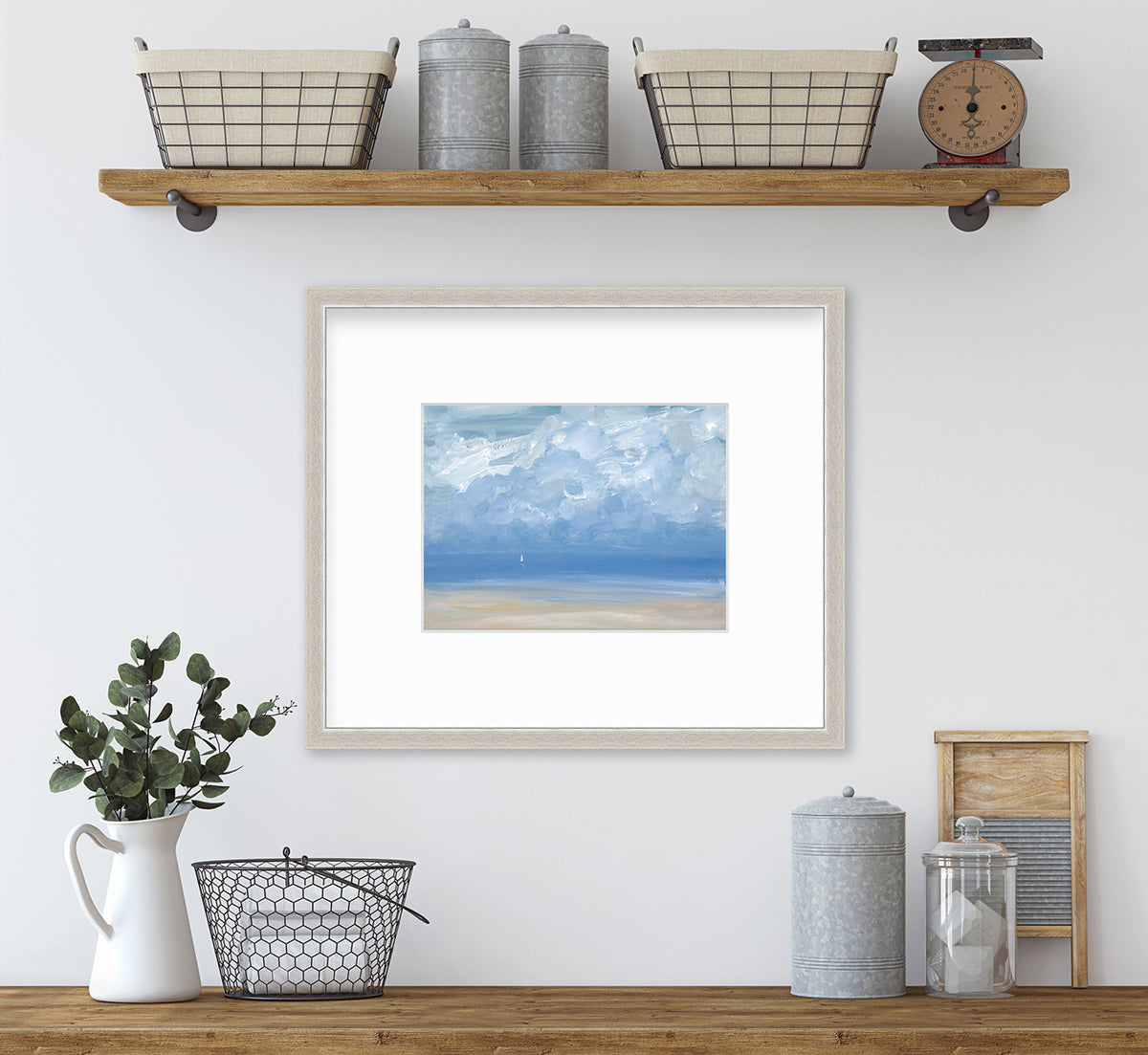 A white, blue and beige abstract seascape print in a silver frame with a mat hangs on a white wall above a wood surface with a black wire basket, a white pitcher with eucalyptus stems and a hammered metal grey cylinder.