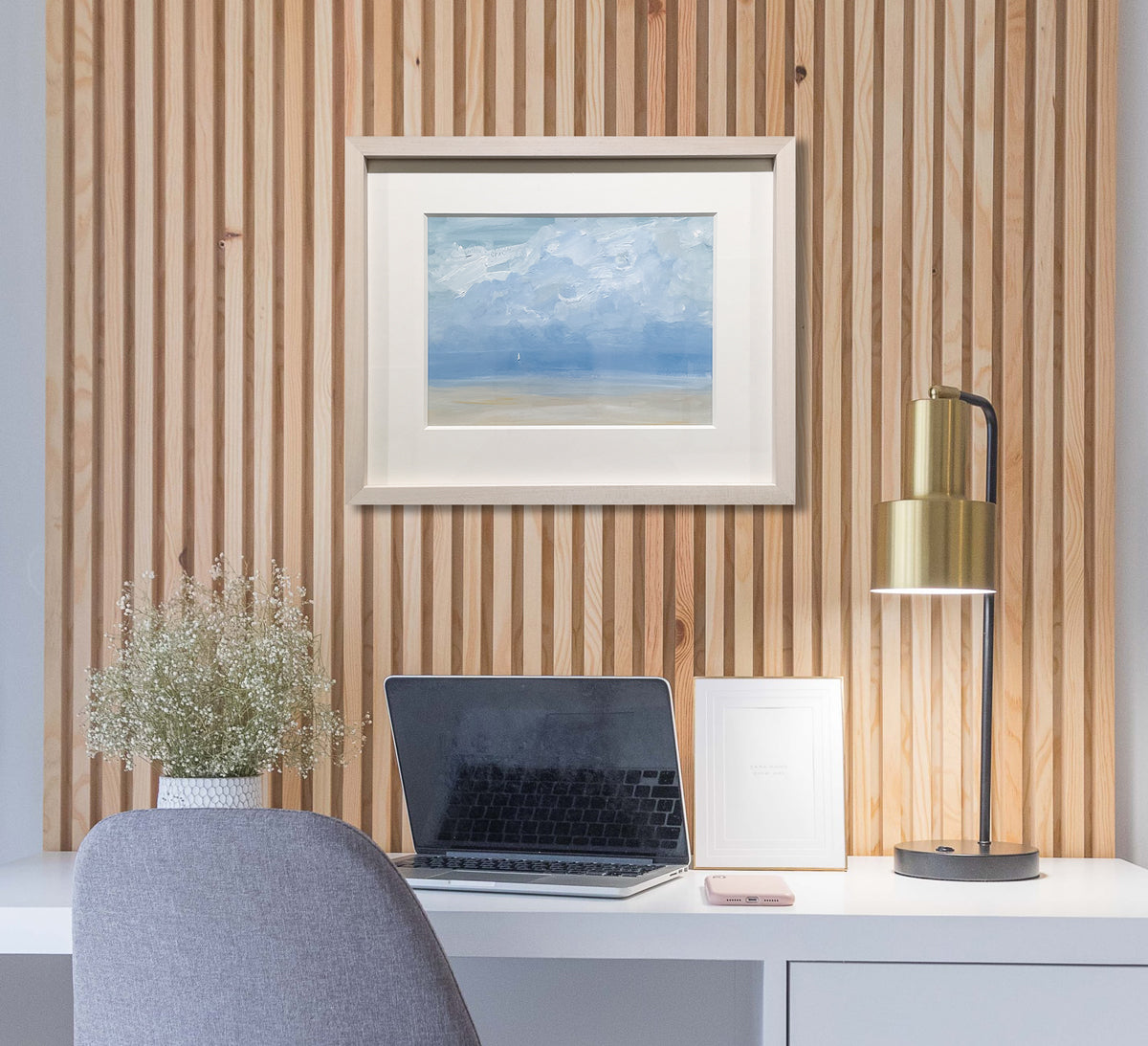 A light blue, white and beige abstract seascape painting with a sailboat by S. Cora Aldo in a natural wood frame with a mat hangs on a wood paneled wall above a white desk with a gold desk lamp, a laptop computer and a potted floral arrangement.